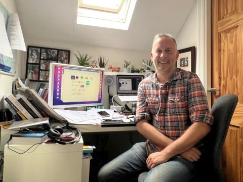 A man sits at a busy desk at home, smiling at the camera