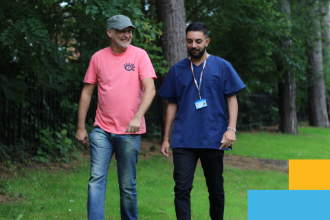 Two people walk in green area by trees, smiling and talking, one wearing casual clothes and the other in medical clothing